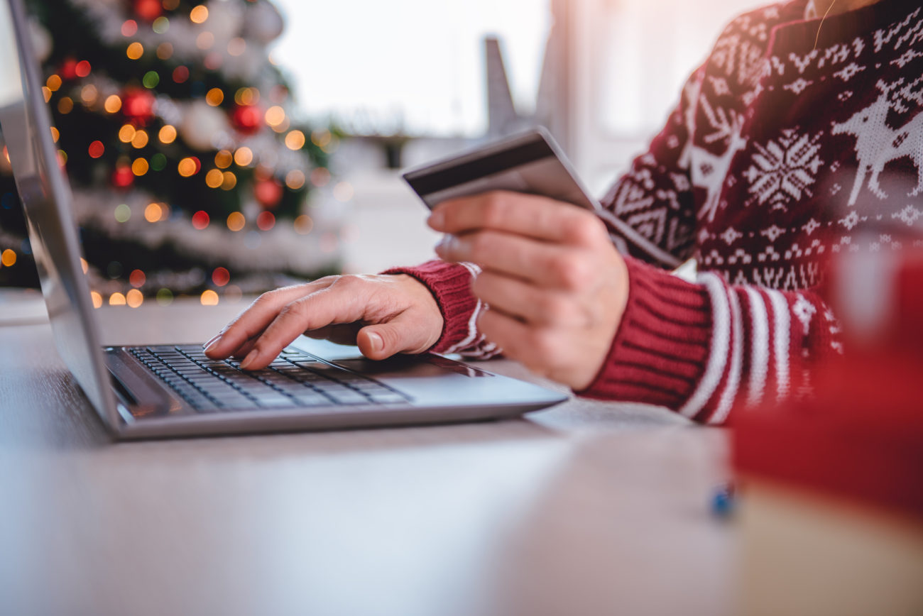 A woman shops online using her laptop and holding her credit card, while a Christmas tree can be seen in the background