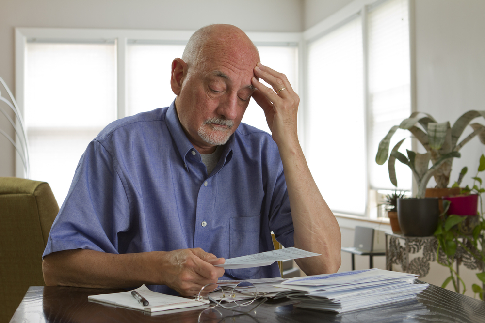 An older man sits at his kitchen table looking at all his bills while scratching the side of his head like he is confused and stressed out