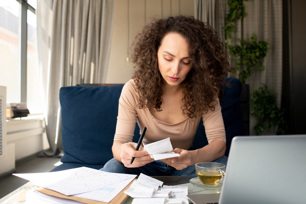 A young woman wears a serious expression on her face as she holds a receipt and adds up her debt