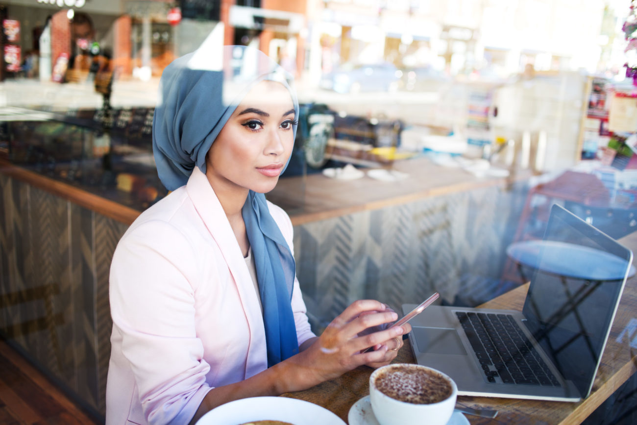 A young woman uses her phone and computer while sitting in a cafe to find a personal loan