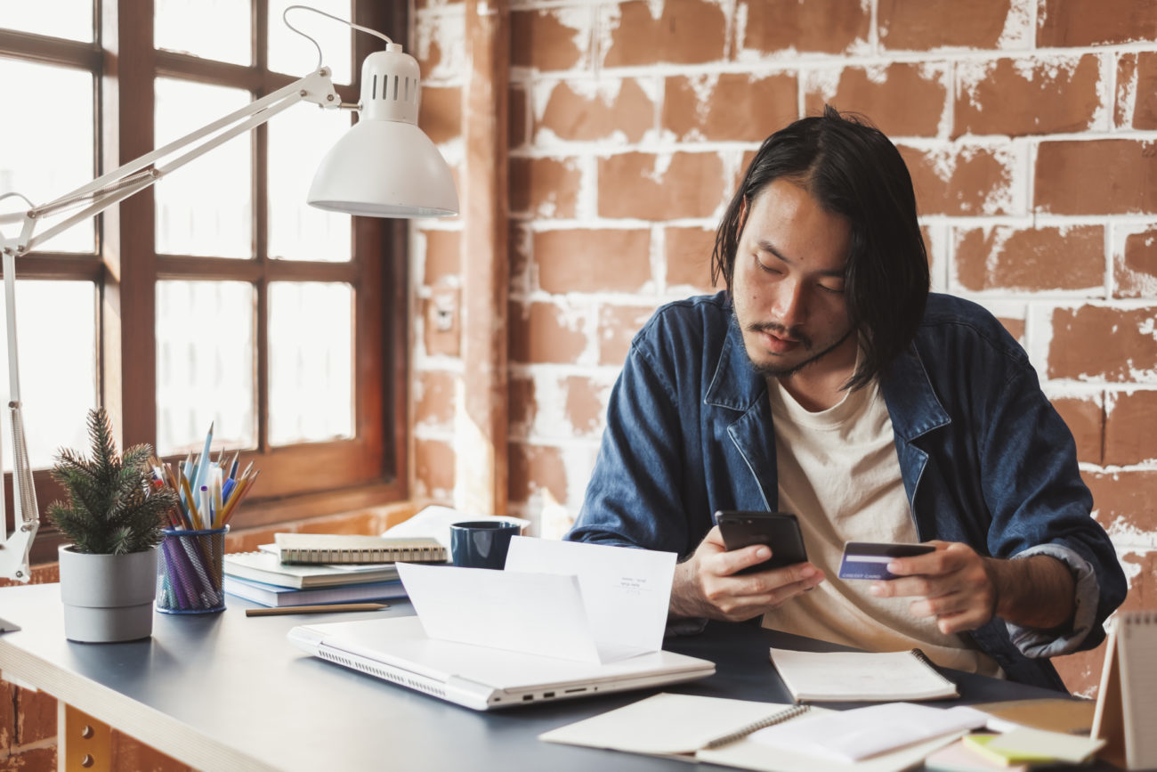 A man sits at a desk with his cell phone in his hand while he works on getting a debt consolidation loan