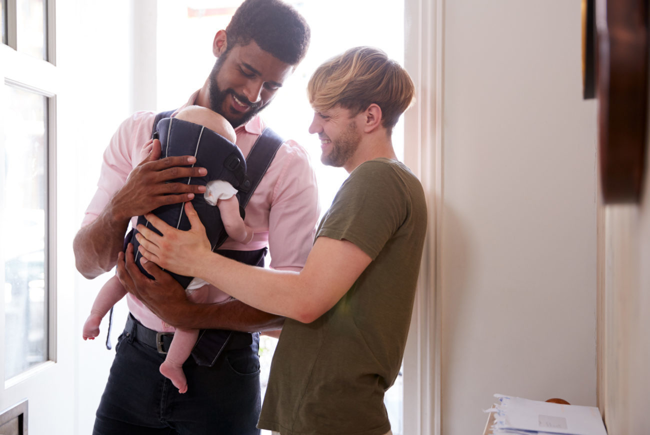 Same Sex Male Couple With Baby Daughter In Sling Opening Front Door Of Home