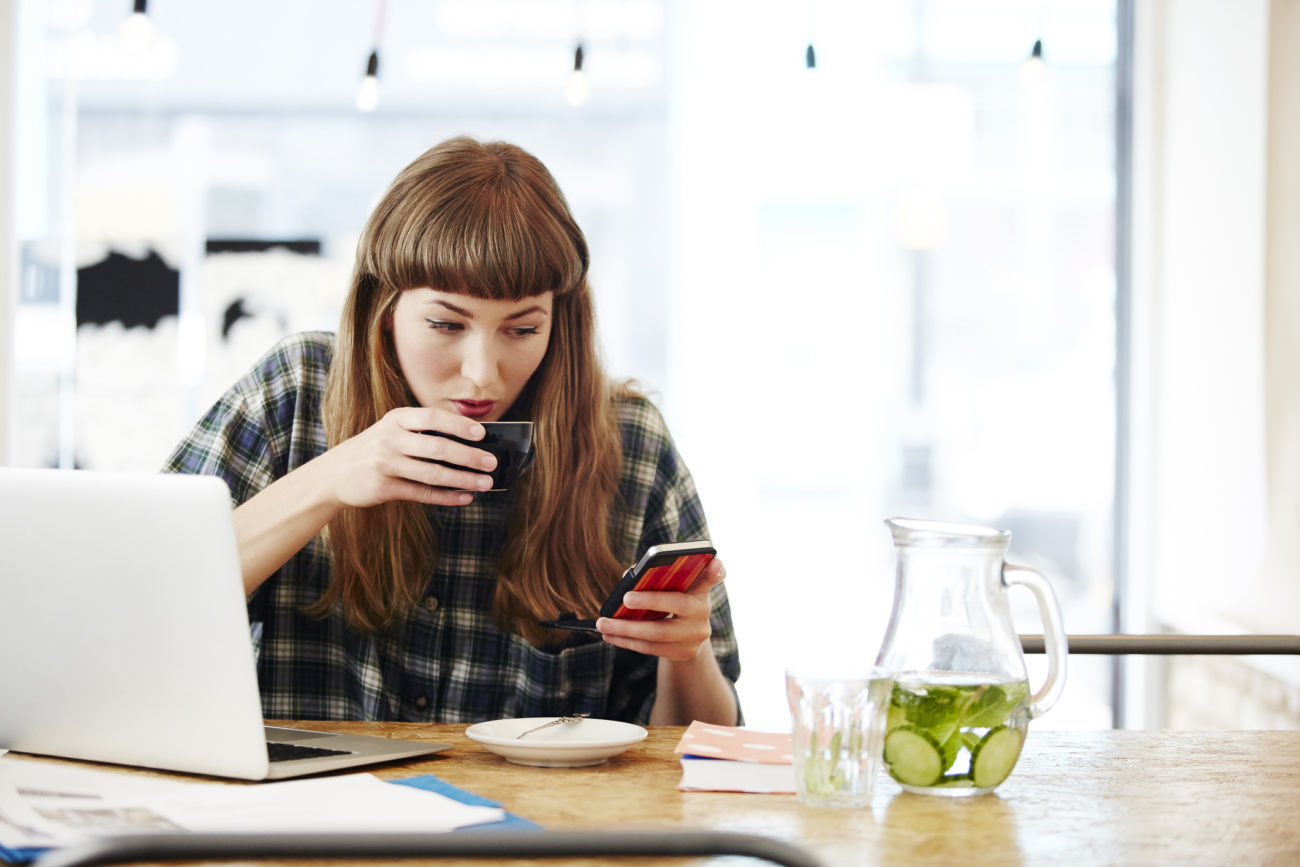 A young woman drinks a cup of coffee while sitting in front of her computer while checking personal loan rates on her phone