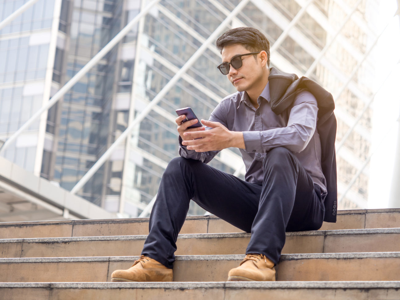A young professional looks at his phone while sitting on stairs outside of a large building