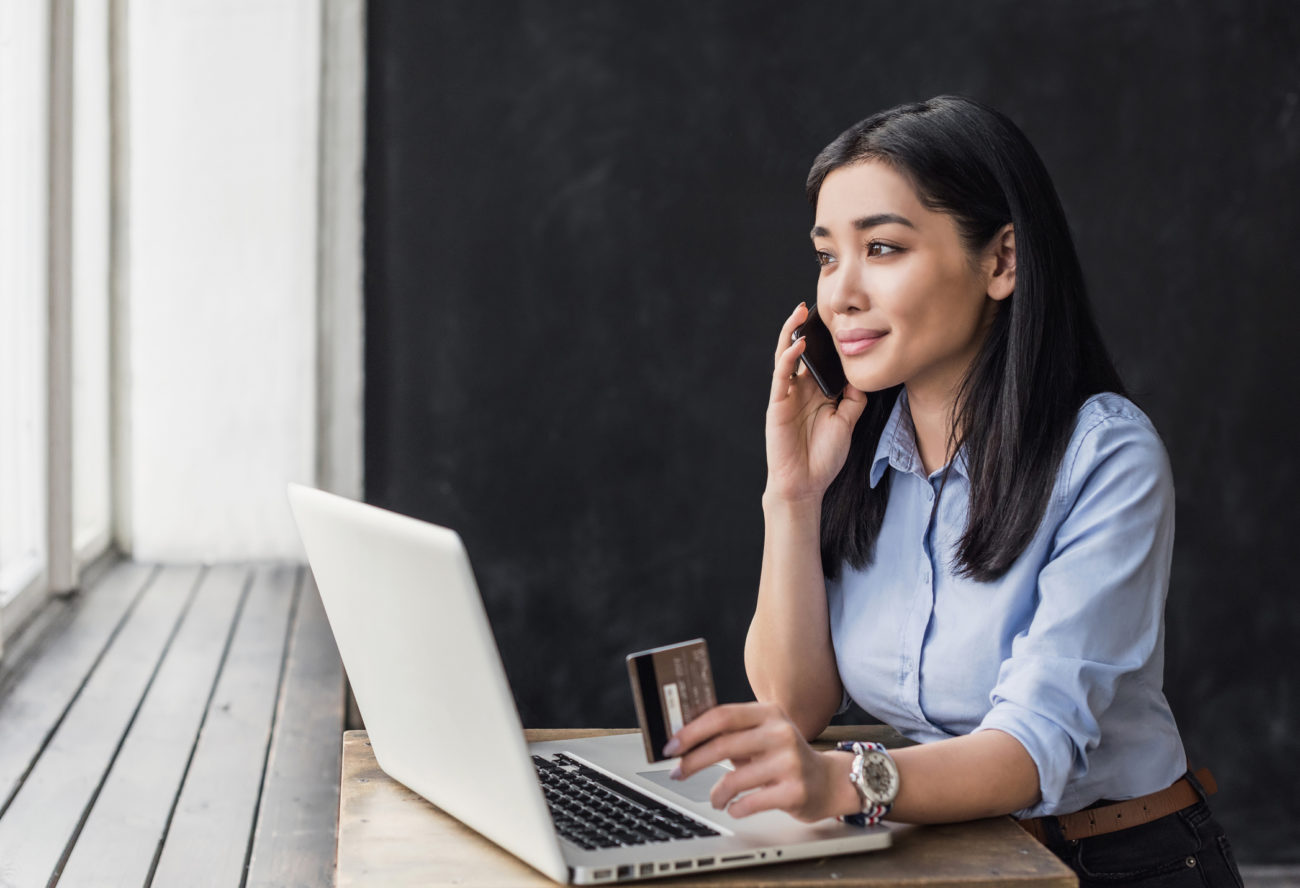 A woman looks at a computer while holding a credit card and talking on the phone.