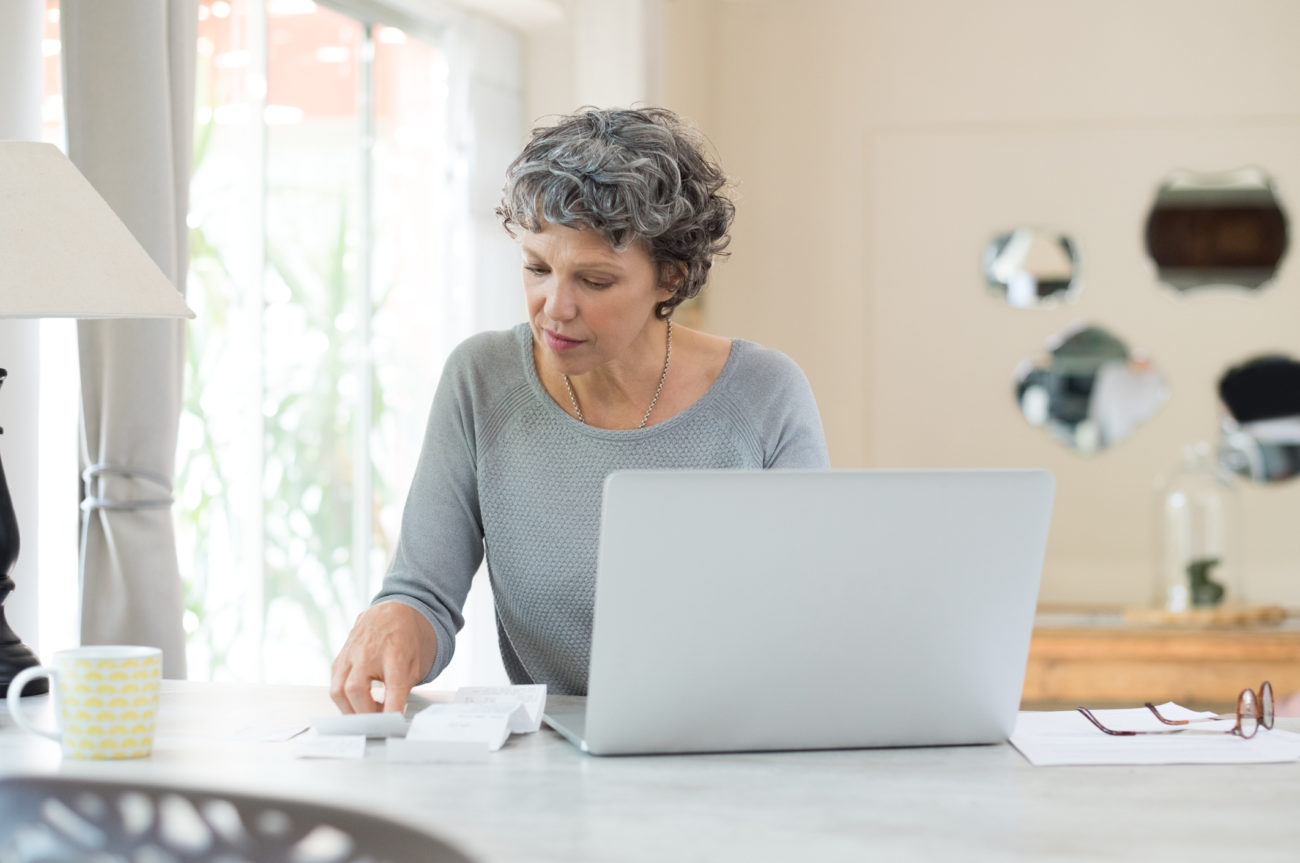 A woman sits at a table with her computer and works on paying household bills