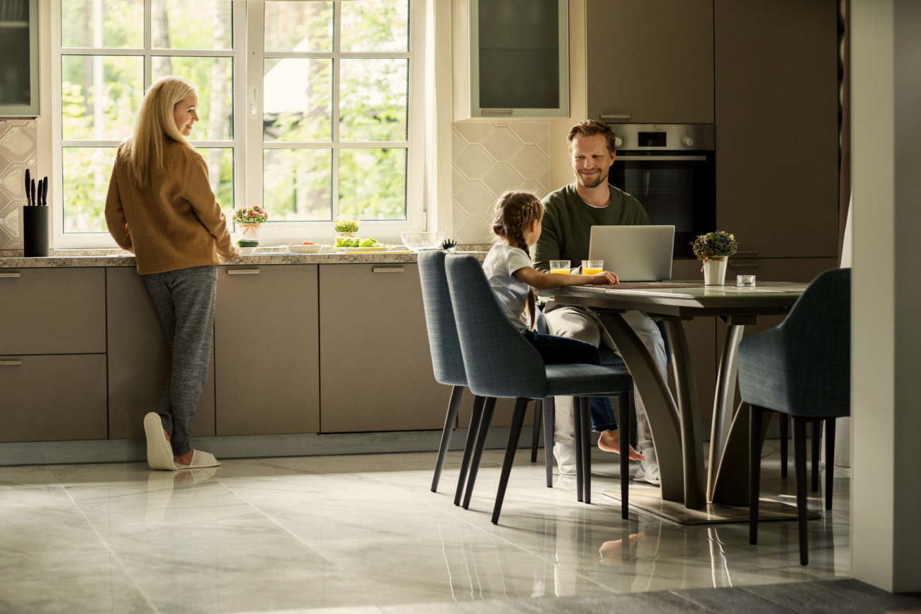 A family gathers in the kitchen while a father sits at the table looking at his computer, and his child sits next to him.