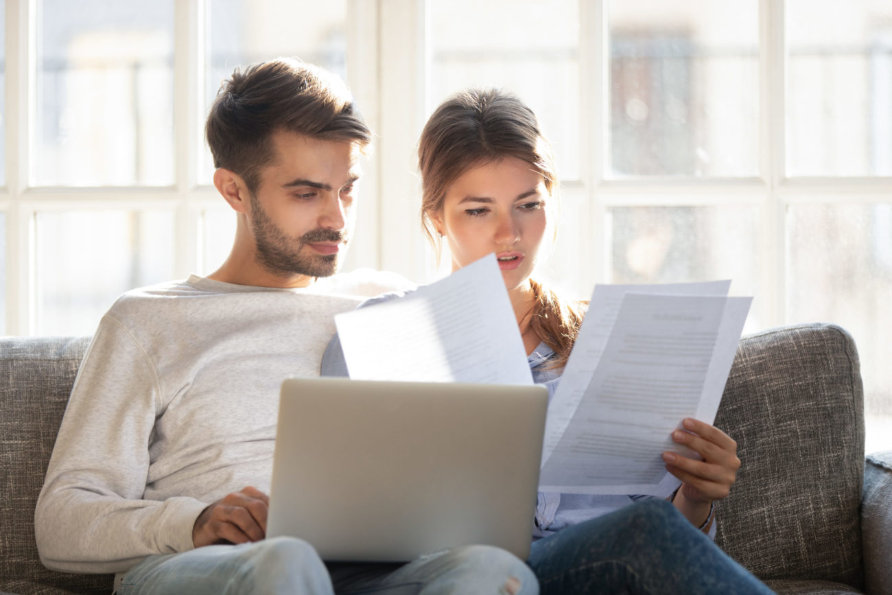 Man and woman sitting on couch with laptop computer looking at paper bills