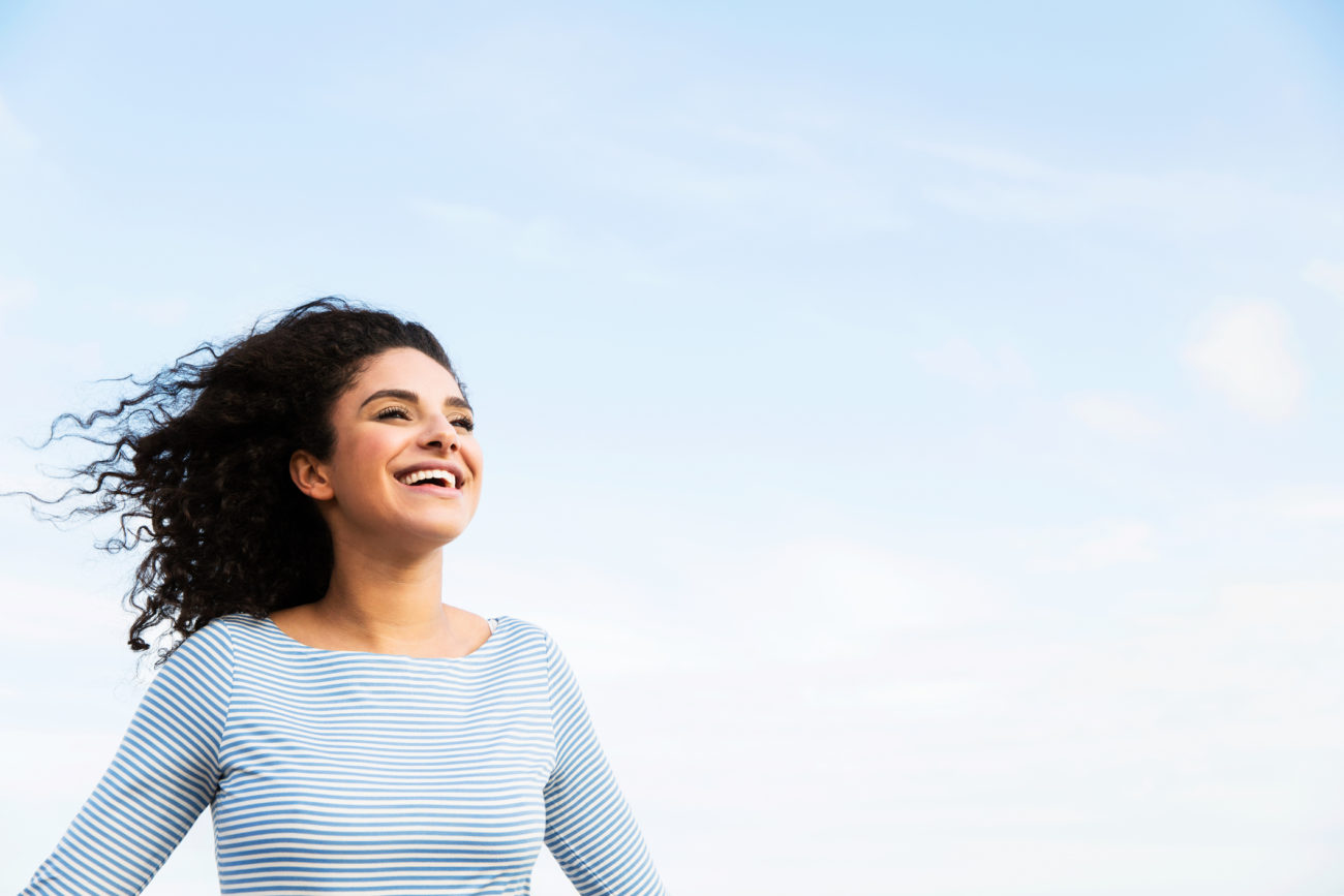 A woman stands outside against a blue sky looking happy and carefree