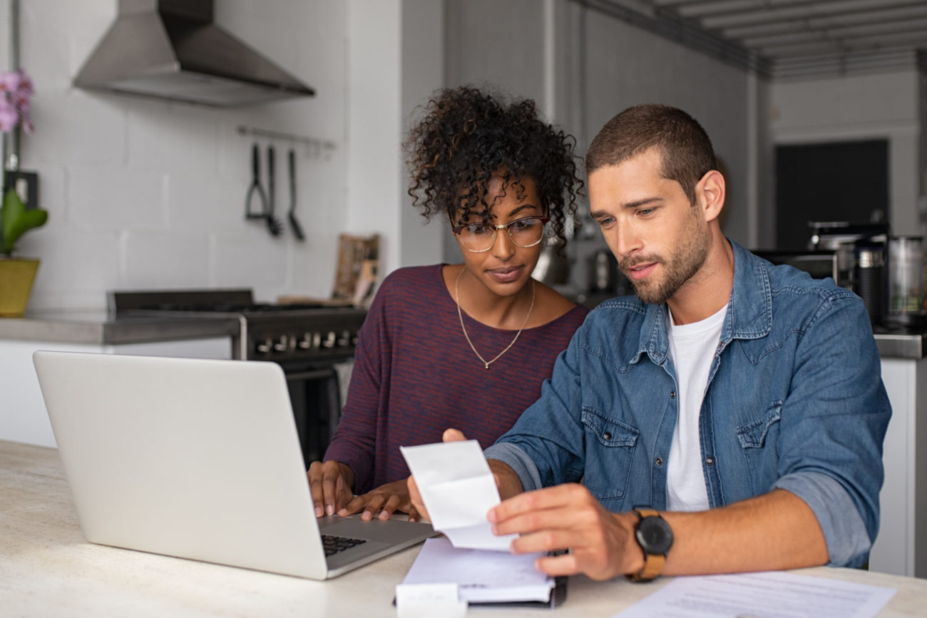 A young couple works on paying their bills while sitting at the kitchen table in front of a laptop computer