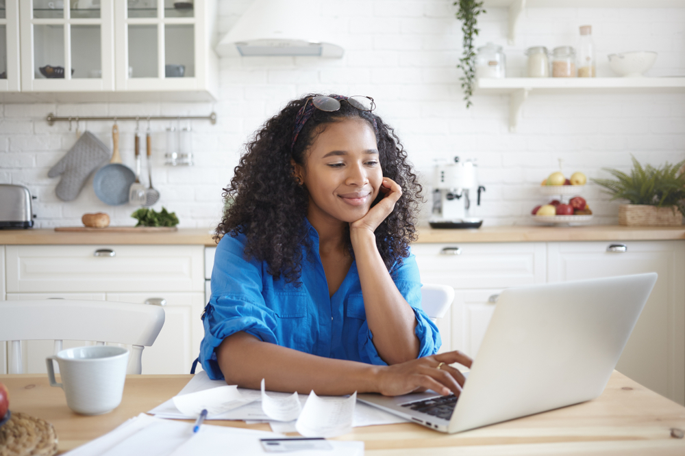 A young woman works on her credit and pays her bills while sitting at a table and looking at her laptop