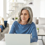 A woman researches personal loan origination fees on her computer while her husband lounges on a couch in the background