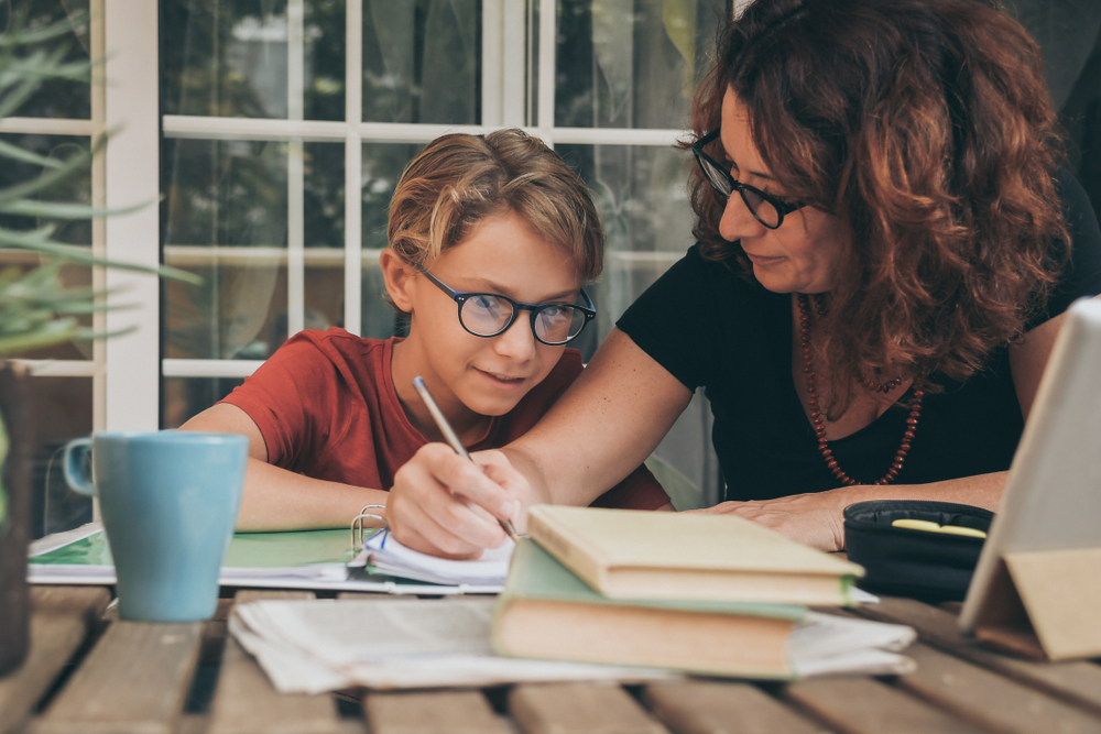 A mother and son sit at a table with a computer and school work spread about as they engage in