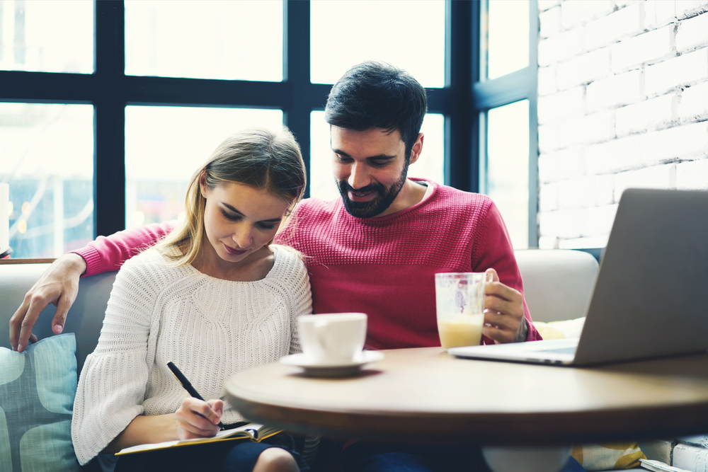 A couple sit in a cafe in front of a laptop computer while the woman takes notes on a piece of paper