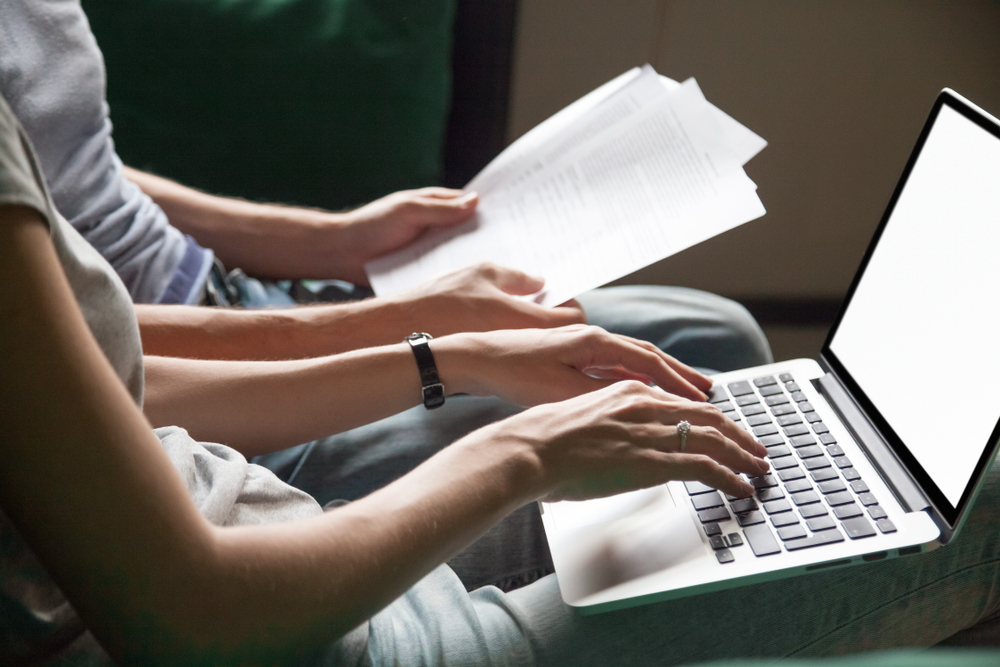 A person types on a laptop while another one holds several papers. They are carefully applying for a loan online