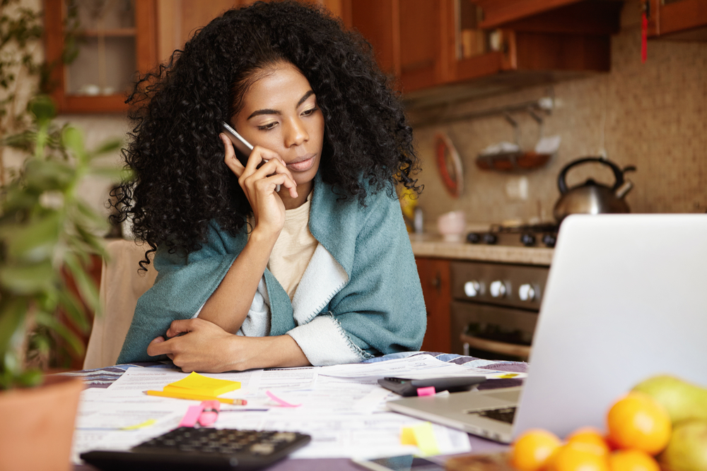 A woman with a concerned expression sits at a table where her monthly bills are spread out. She talks on the phone and looks at her computer.