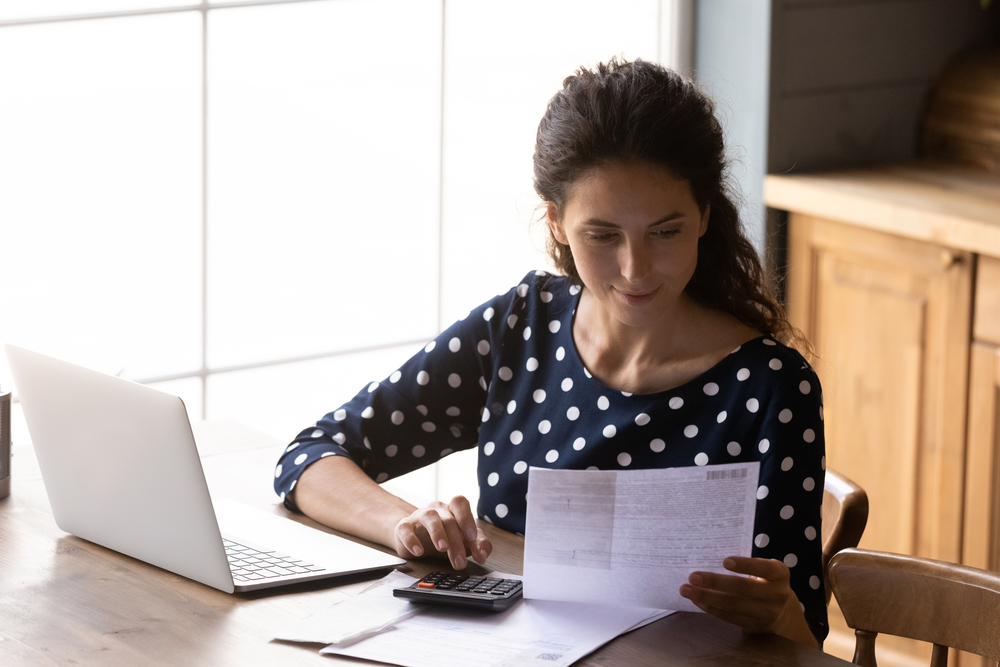 A woman works on her tax return while she sits at the table in her kitchen using a calculator