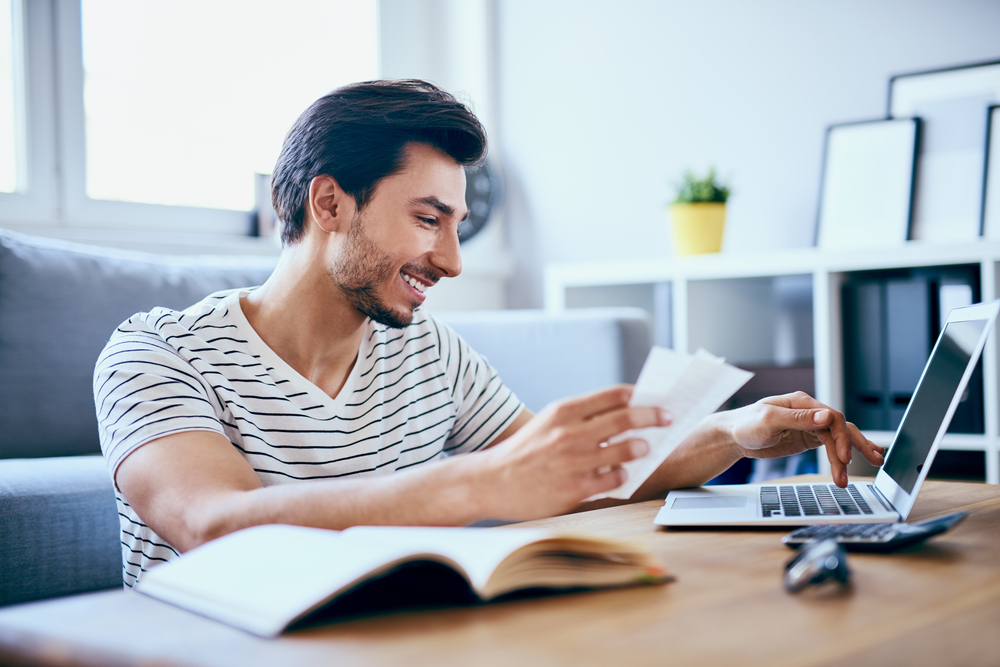 A man is sitting at a table with his computer in front of him as he holds a piece of paper and pays his bills online