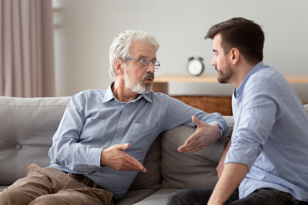 An older man has a discussion with adult son about lending him money as they sit on a couch and face each other