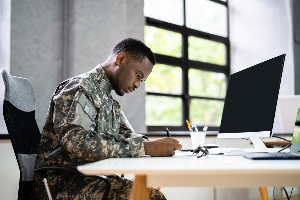 A military member wearing a uniform fills out a form as he sits at a desk in front of a computer